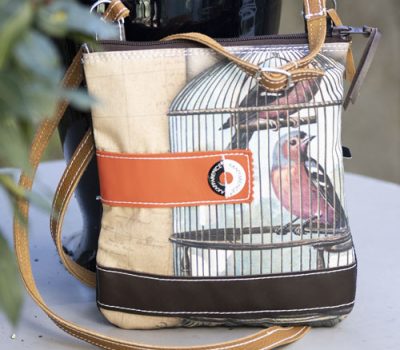 pocket en tissus beige avec une illustration de 2 oiseaux dans une cage avec une bande en cuir orange qui s'arrête au milieu du sac et le logo cousu dessus et une bande en cuir noir en dessous du sac